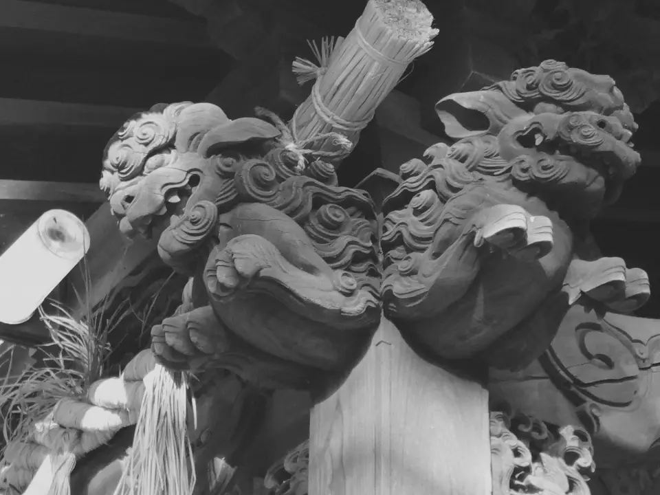 Carved wooden komainu guardians and a shimenawa rope under a shrine roof. Black-and-white photo.
