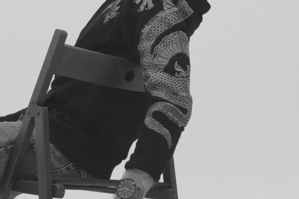 A black hoodie sleeve with an embroidered dragon, a wristwatch and a wooden chair. Black-and-white photo.